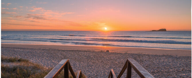 Sunrise over a sandy beach with calm ocean waves and a small island in the distance.
