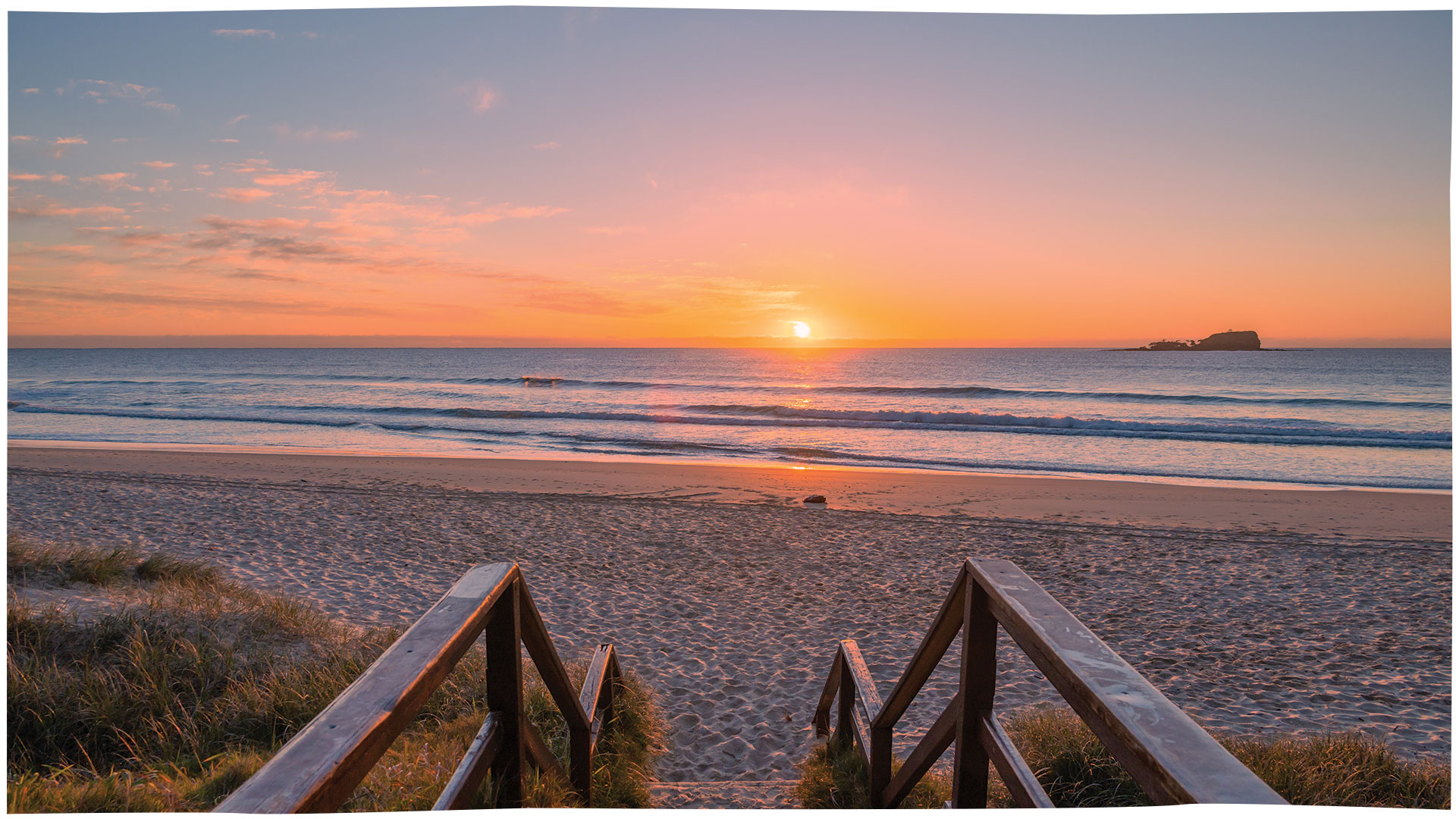 Sunrise over a sandy beach with calm ocean waves and a small island in the distance.